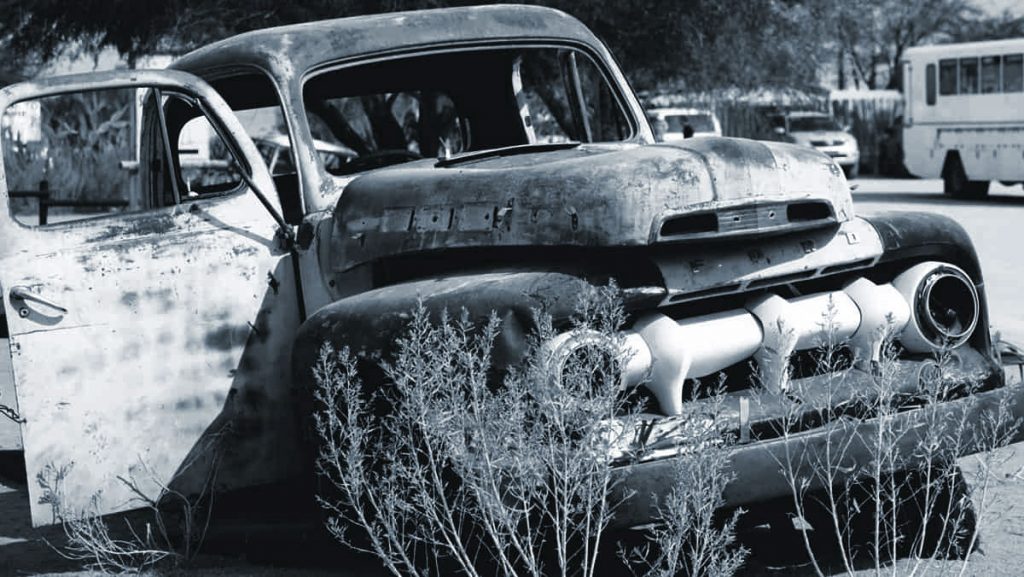 A vintage, rusted car with an open door surrounded by dry grass, with an old bus in the background, reflecting a nostalgic scene.